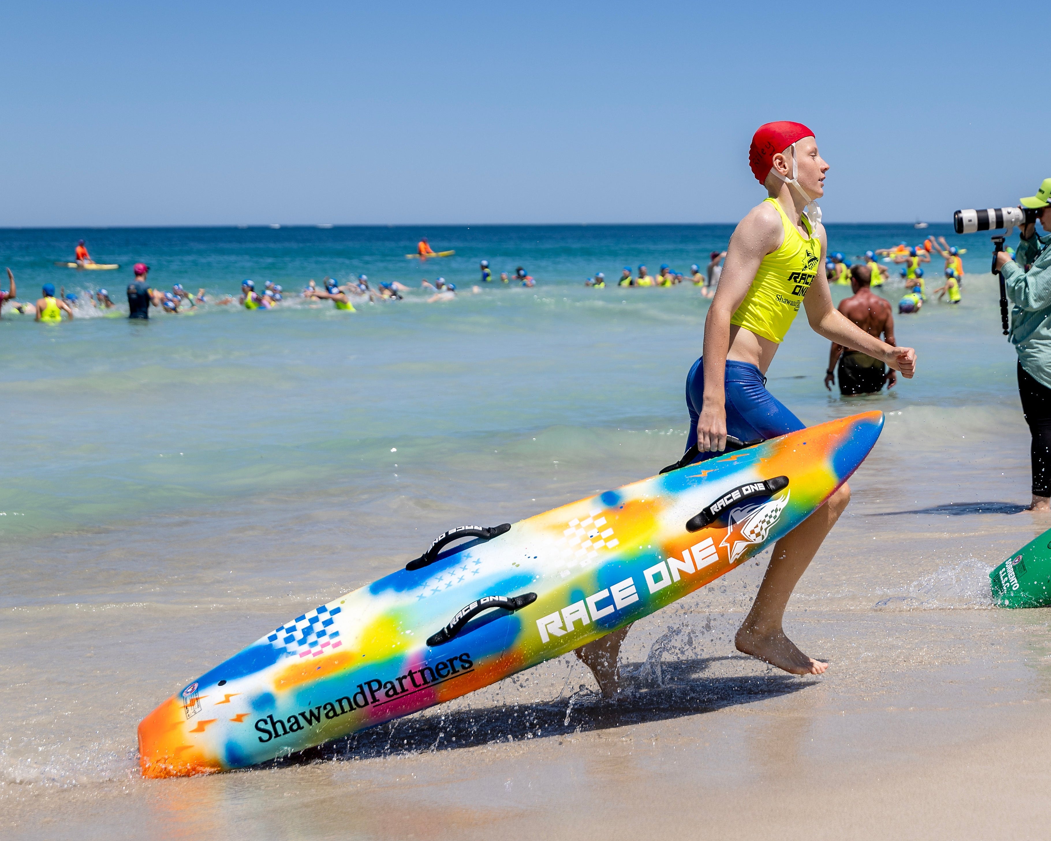 Two people carrying surfboards on a beach with a clear blue sky.
